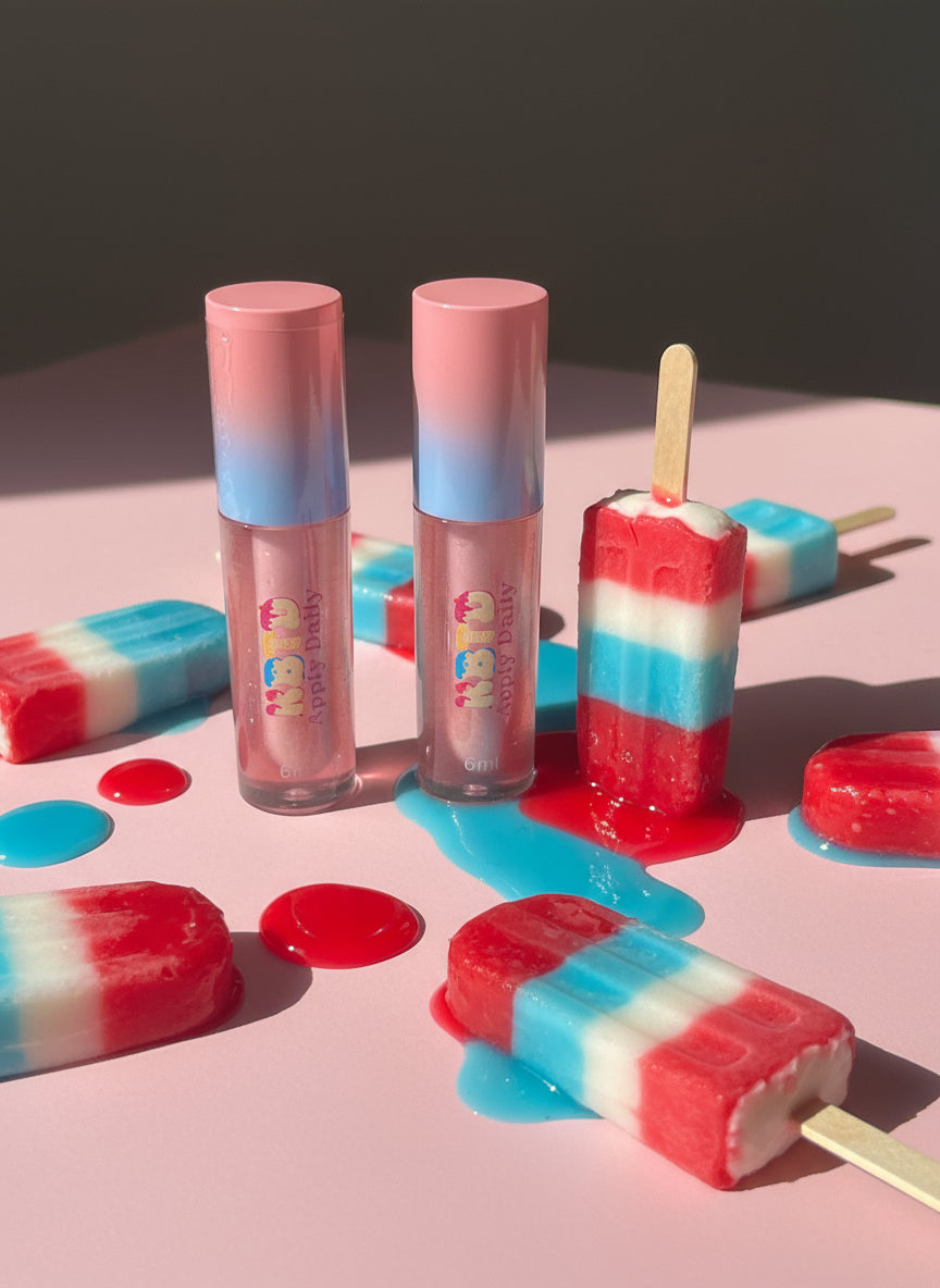 Lip gloss tubes with pink/blue ombré cap and popsicle-shaped candies on a pink surface with red, white, and blue nostalgic colors. popsicle flavored.